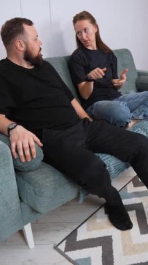 A man and a woman are sitting on a comfortable sofa, having a lively conversation, gesturing to emphasize their points of view in the cozy atmosphere of the living room. vertical video