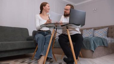 Business partners working from home are sitting at a glass table, woman holding documents and discussing with a man looking at laptop, having a video call