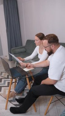 Two colleagues, a man and a woman, are seated at a glass table, intently focused on a laptop screen and discussing documents, engaging in collaborative problem-solving within a shared office space