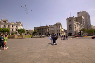 Lima, Peru - 18 Nisan 2018: Arka planda Hostal San Martin de dahil olmak üzere bahçeleri ve binaları olan San Martin Meydanı 'nda yürüyen ve konuşan turistler
