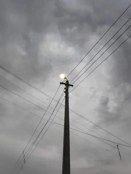 Bulutlu havada ampullü lamba direği ve yanında ve üstünde elektrik kabloları var.