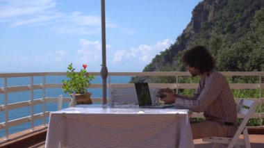 Kıvırcık adam balkonda deniz manzaralı dizüstü bilgisayarla çalışıyor.