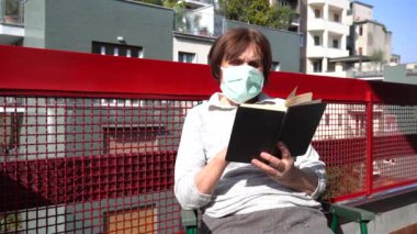 Avrupa, İtalya, Milano - 70 yaşındaki karantina maskeli kadın kendini Coronavirus 19 salgınından korumak için çeşitli iç faaliyetlerde bulunuyor - bir kitap oku