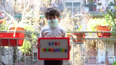 Maskeli çocuk karantina sırasında elinde Coronavirus posteriyle balkonda oturuyor.