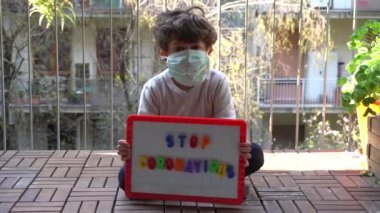 Maskeli çocuk karantina sırasında elinde Coronavirus posteriyle balkonda oturuyor.