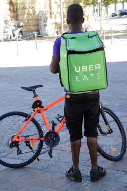 Şehirdeki nehrin önünde bisikletli bisikletçinin arka manzarası, Uber yemek yiyor.
