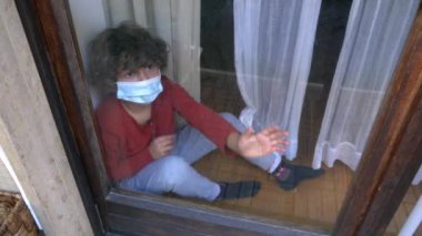 Avrupa, Amerika ve Asya 'daki Coronavirus tecridi sırasında 6 yaşında beyaz beyaz beyaz beyaz bir çocuk maske takmış karantina evinde evin penceresinden dışarı bakıyor.