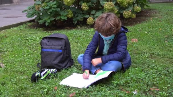 可爱的男孩在户外坐在草地上做作业 戴着口罩 — 图库视频影像