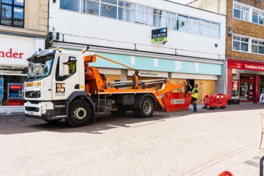 A skip lorry by a shop under refurbishment, with the driver using controls to lift a skip of building waste onto the lorry, in Trowbridge Wiltshire clipart