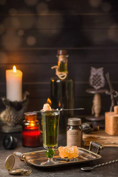 Absinthe poured into a tall glass. On top is a spoon with burning sugar. In the background is a bottle of absinthe. On a black wooden background with burning candles. Romantic style.