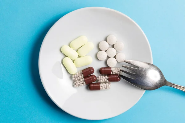 Vitaminas y medicamentos en forma de varias píldoras y píldoras en un plato blanco con un tenedor sobre un fondo azul . — Foto de Stock