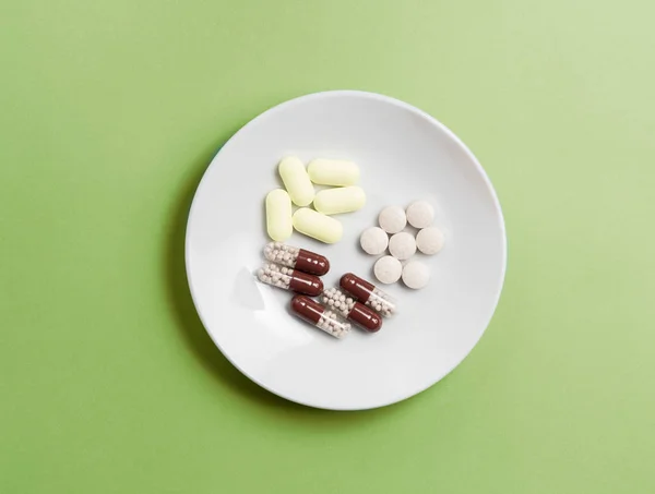 Vitaminas y medicamentos en forma de varias píldoras y píldoras en un plato blanco sobre un fondo verde . — Foto de Stock