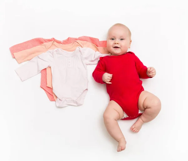 Un bebé feliz de cuatro meses en traje de cuerpo rojo acostado sobre un fondo blanco con ropa rosa de menor tamaño . —  Fotos de Stock