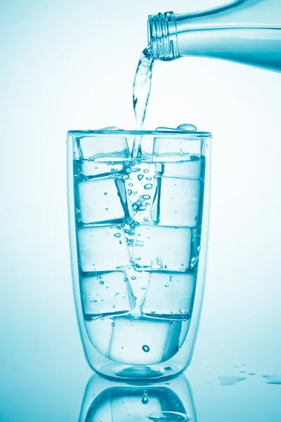 Despeje água de uma garrafa de vidro em vidro com cubos de gelo no fundo azul — Fotografia de Stock