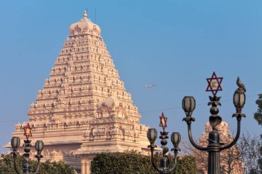 View of lamps with a traditional Hindu six-pointed star, and the main building of the Chattarpura temple complex near by which the passenger plane flies by. clipart
