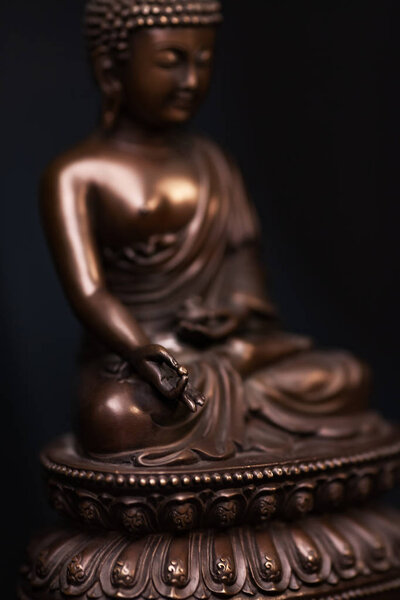 Defocused silhouette of Buddha's figure, brown color made of metal in a meditation pose with the hands keep a pearl of knowledge.