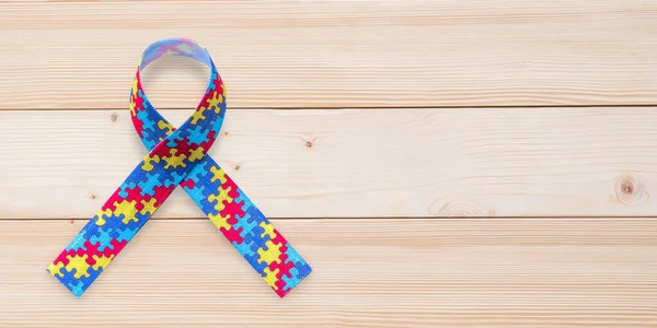 Autism awareness ribbon in puzzle or jigsaw pattern (isolated on wood background with clipping path) for World Autism Awareness day, mental health care concept for autistic child person support and family nursing care