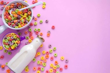 Una taza grande blanca y una ms pequea de color lila, ambas llenas de aros de tahıl. Cerca a ellas una botella de leche fresca.