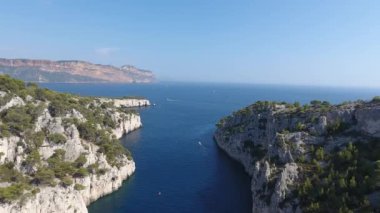 İnsansız hava aracı akdeniz üzerinde uçuyor, dereden iniyor. Güneşli bir gün, kayalıklar ve ağaçlar. Arka planda Cassis