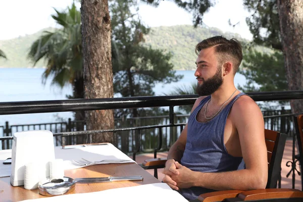 Young man with black hair and beard sitting in a restaurant in the Park in a summer evening and green trees arround