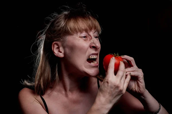 Middle-aged woman holding an tomato in her hands and she does not like it. Denial of a healthy lifestyle. Photoshoot in dark key