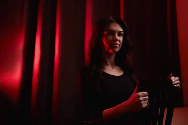 Mysterious portrait of a girl in black dress and red light background. Actress before going on stage. Photo shoot in a dark key