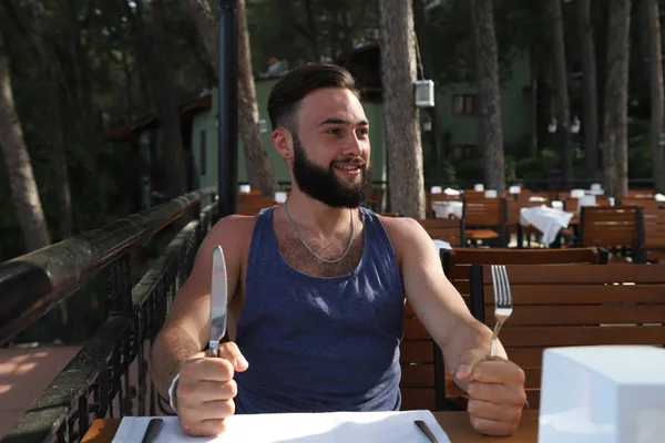 Young man with black hair and beard sitting in a restaurant in the Park in a summer evening and green trees arround