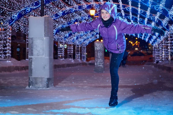 Girl skating on the ice arena full of light on the city square in winter evening. Photo shoot with a teenager female athlete with blue lights on a snowy winter night