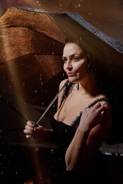 Beautiful sexy girl in black with umbrella and water spray in the Studio on a dark background with orange back light