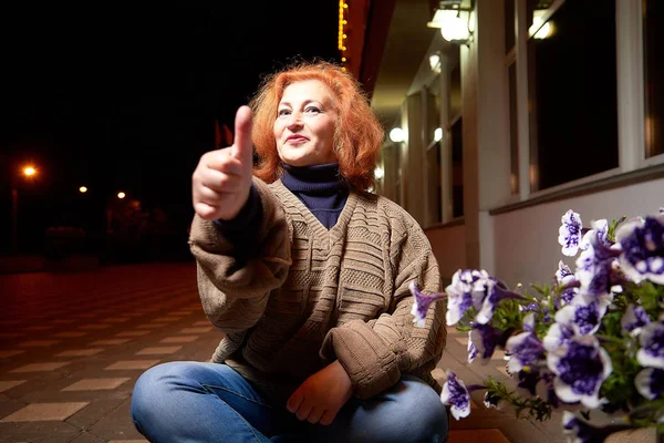 Emotional middle-aged woman in a thick gray sweater on a city street at night. Elderly female pensioner in the evening and darkness behind her