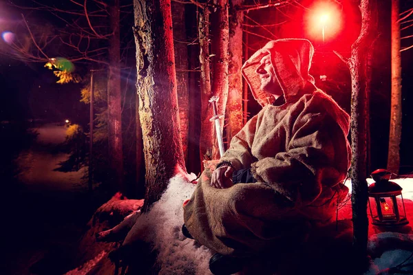 Medieval monk in canvas sackcloth robe praying in dark forest with snow and red light on winter night. Fantasy or fairy tale about wandering monk. Story about the forces of good and evil in the world
