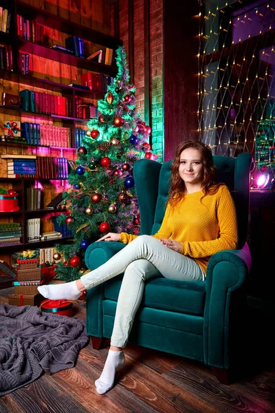 A girl in white jeans, a yellow sweater sitting in a chair in a room with a Christmas tree prepared for Christmas or New year. Interior of a private room before the holiday