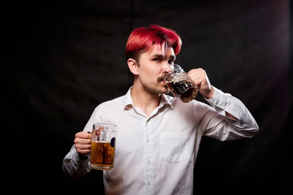 Young handsome guy with red hair in white shirt drinking beer. Funny man with emotions on the face and beer mug in hand and black background. Alcoholic is happy with alcohol. Model in studio