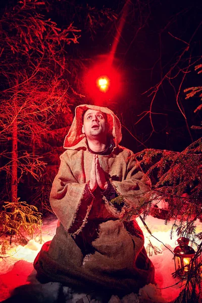 Medieval monk in canvas sackcloth robe praying in dark forest with snow and red light on winter night. Fantasy or fairy tale about wandering monk. Story about the forces of good and evil in the world