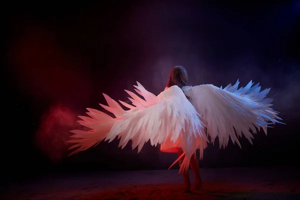 Small young model girl with white wings posing and dancing in dark black studo during photoshoot with flour or dust and colour light. Actress in performance about struggle between good and evil