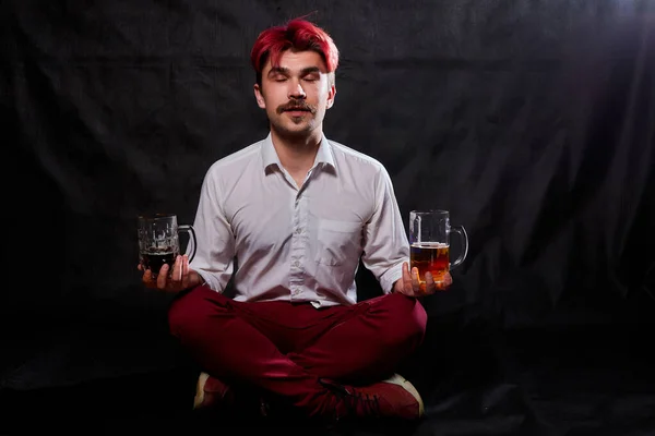 Young handsome guy with red hair in white shirt drinking beer. Funny man with emotions on the face and two beer mugs in hands and black background. Alcoholic is happy with alcohol. Model in studio