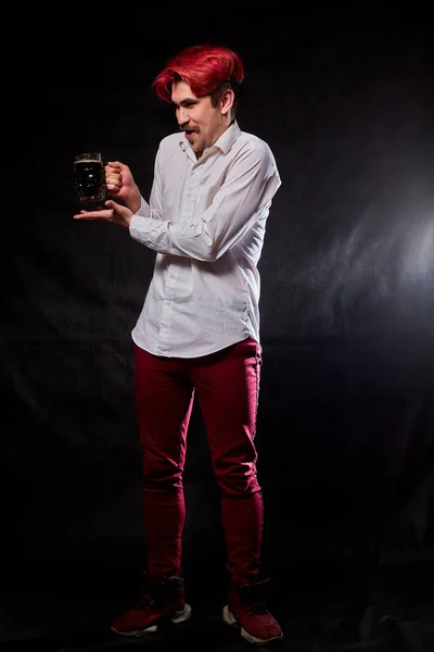 Young handsome guy with red hair in white shirt drinking beer. Funny man with emotions on the face and beer mug in hand and black background. Alcoholic having fun with alcohol. Model posing in studio