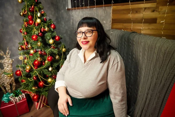 Fat cute woman with glasses and black hair posing in a room decorated for Christmas or New year in Russia