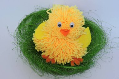 Sarı tüylü, el yapımı, turuncu gagalı, sarı kanatlı Paskalya tavuğu sisal liflerden yapılmış yeşil bir yuvada oturan sarı iplikten yapılmış bir civciv, beyaz arka plan.