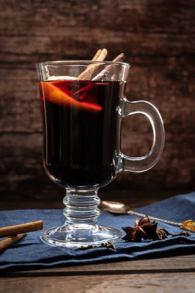 A glass of hot mulled wine with cinnamon sticks and a slice of lemon, with dark blue textile on a dark vintage wooden background — Stock Photo, Image