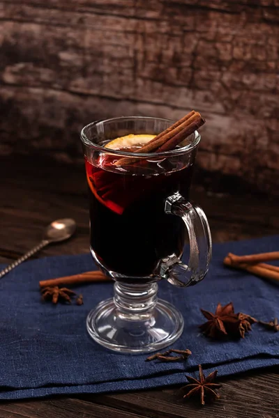 A glass of hot mulled wine with cinnamon sticks and a slice of lemon, with dark blue textile on a dark vintage wooden background — Stock Photo, Image