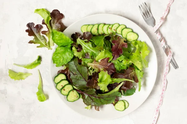 A plate with green salad leaves, sesame seeds and cucumber slices, a tape-measure wrapped around a fork on the right, on a light grey background — Stock Photo, Image