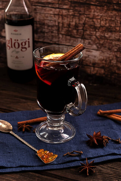 A glass of hot mulled wine with cinnamon sticks and a slice of lemon, with a bottle of Scandinavian "Glogi" dark blue textile on a dark vintage wooden background