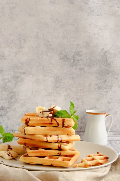 Hot, fresh stack of waffles with banana and chocolate sauce and a white milk jug on grey background with copy space — Stock Photo, Image