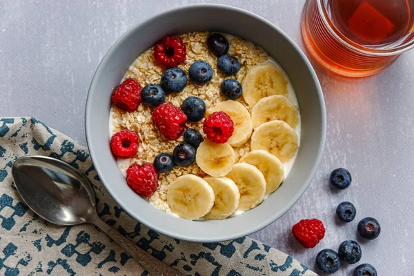 Top view of a healthy breakfast with oatmeal in a bowl, banana slices, raspberry, blueberry and a cup of tea on light grey tabletop — Stock Photo, Image