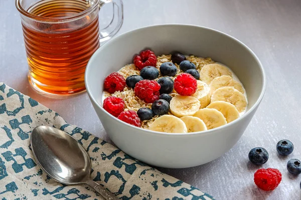 Oldalnézet egy egészséges reggeli zabpehellyel egy tálban, banánszeletek, málna, áfonya és egy csésze tea világosszürke asztalon — Stock Fotó