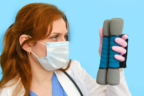 Woman doctor holds dumbbells for sports on a blue background, close-up.