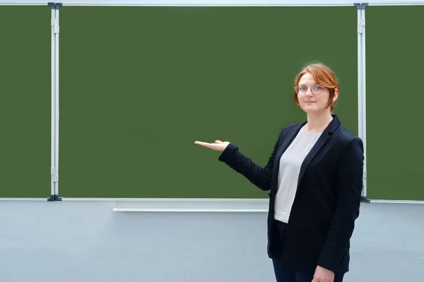 Woman teacher smiles and points to an empty blackboard, copy space