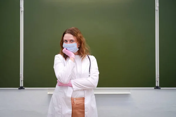 A school doctor in a medical mask stands thinking at the blackboard, copy space