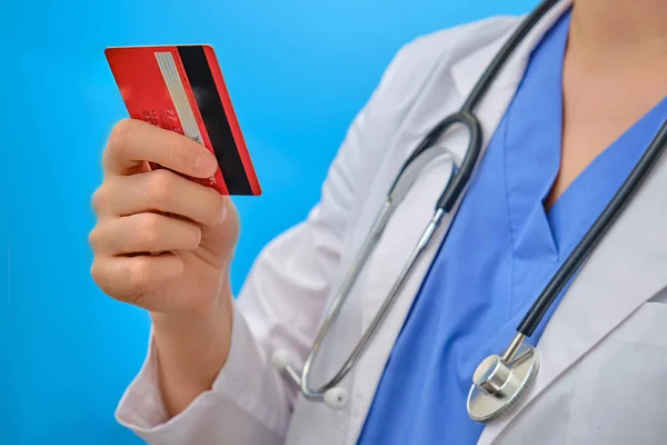 The doctor is holding a plastic card, close-up on a blue background. Woman nurse with a red bank card. Concept of the financial crisis due to the coronavirus pandemic in the world